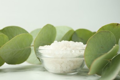 Photo of Sea salt in glass bowl and eucalyptus leaves on white marble table, closeup