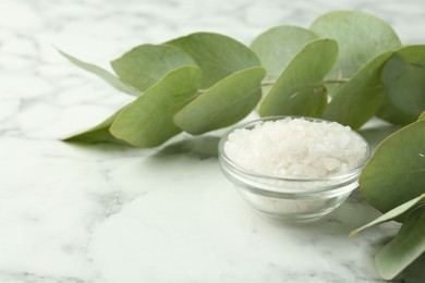 Photo of Sea salt in glass bowl and eucalyptus leaves on white marble table, closeup