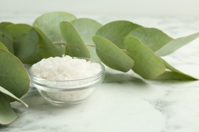 Sea salt in glass bowl and eucalyptus leaves on white marble table, closeup