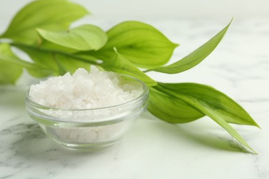 Photo of Sea salt in glass bowl and green leaves on white marble table, closeup