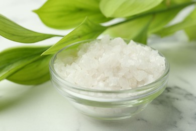 Photo of Sea salt in glass bowl and green leaves on white marble table, closeup