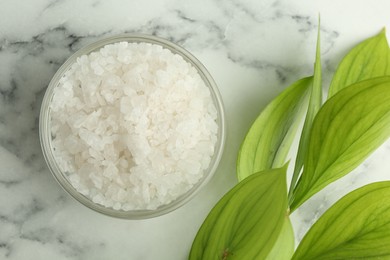 Photo of Sea salt in glass bowl and green leaves on white marble table, top view