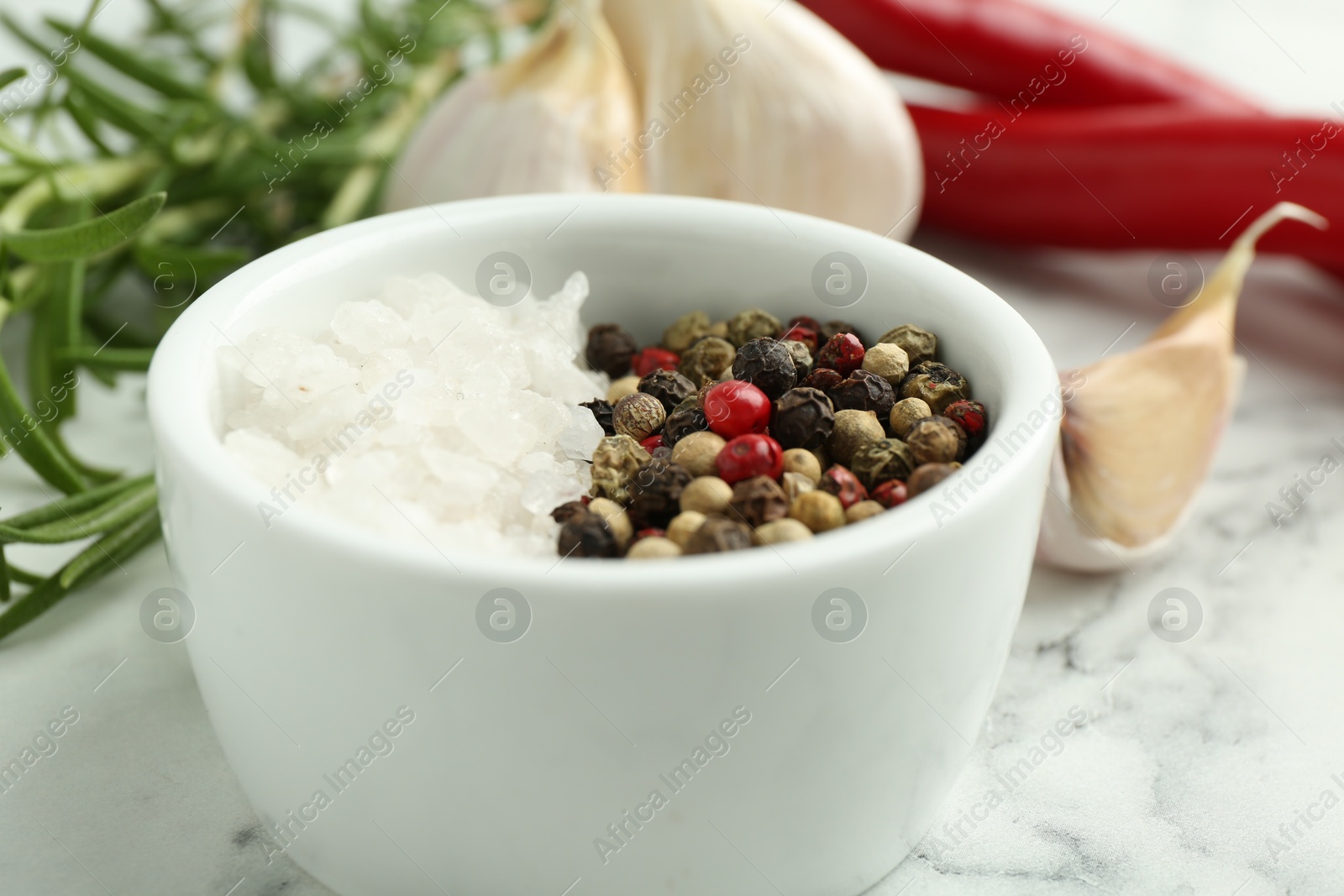 Photo of Sea salt, peppercorns, rosemary, chili peppers and garlic on white marble table, closeup