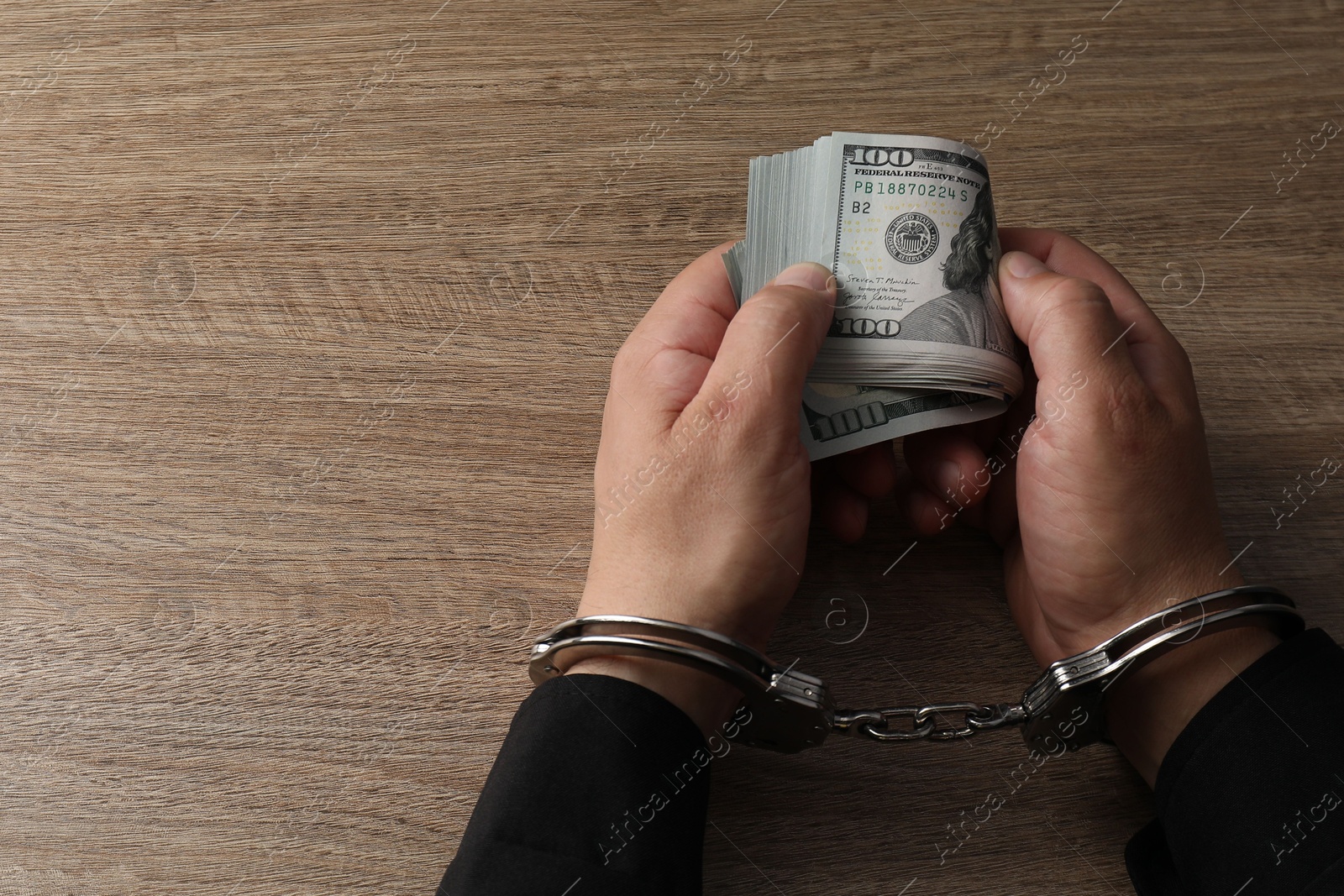 Photo of Corruption. Man in handcuffs holding dollar banknotes at wooden table, closeup