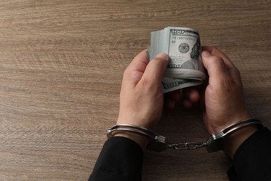 Corruption. Man in handcuffs holding dollar banknotes at wooden table, closeup