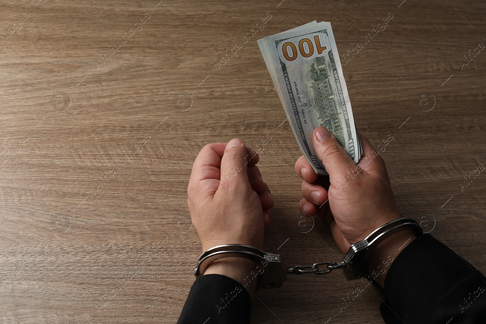 Photo of Corruption. Man in handcuffs holding dollar banknotes at wooden table, closeup