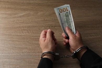 Corruption. Man in handcuffs holding dollar banknotes at wooden table, closeup