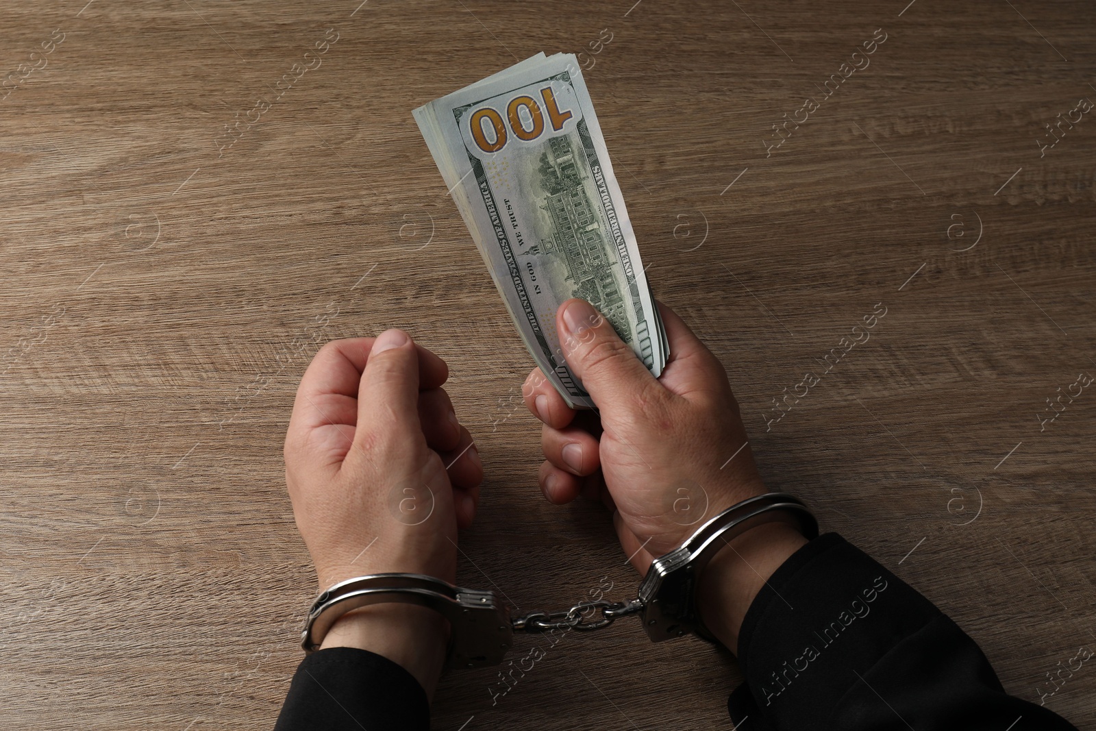 Photo of Corruption. Man in handcuffs holding dollar banknotes at wooden table, closeup