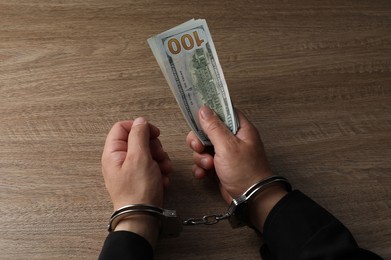 Corruption. Man in handcuffs holding dollar banknotes at wooden table, closeup