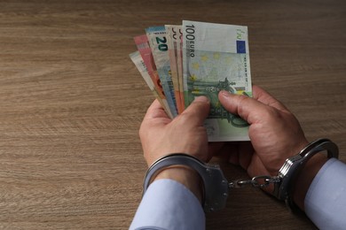 Corruption. Man in handcuffs holding euro banknotes at wooden table, closeup