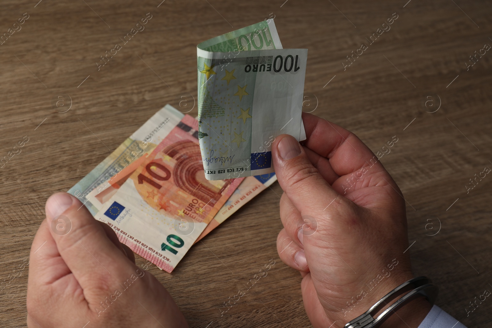 Photo of Corruption. Man in handcuffs holding euro banknotes at wooden table, closeup