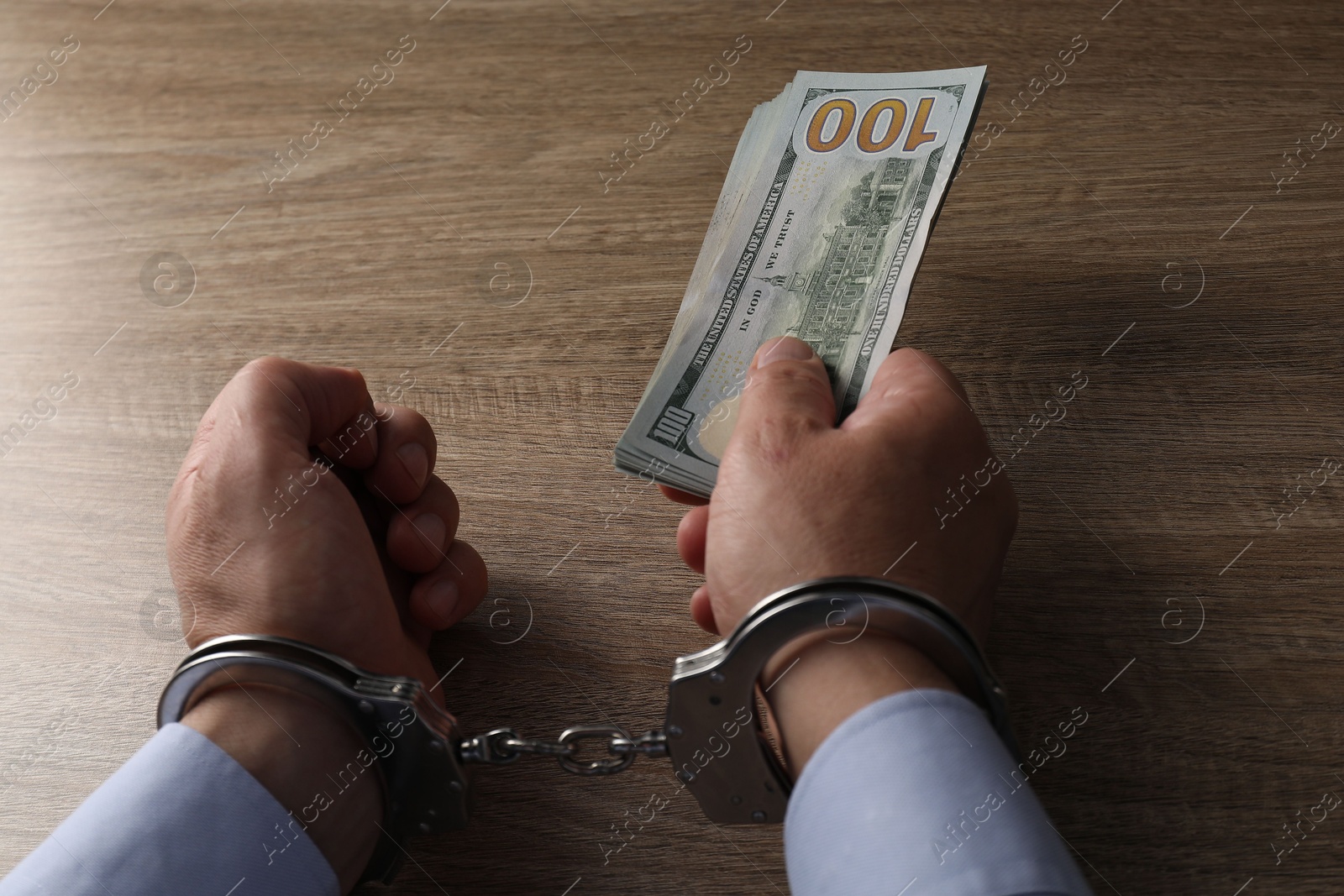 Photo of Corruption. Man in handcuffs holding dollar banknotes at wooden table, closeup