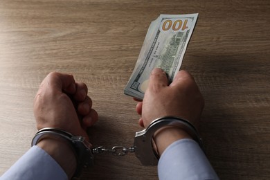 Photo of Corruption. Man in handcuffs holding dollar banknotes at wooden table, closeup