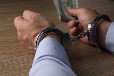 Photo of Corruption. Man in handcuffs holding dollar banknotes at wooden table, closeup