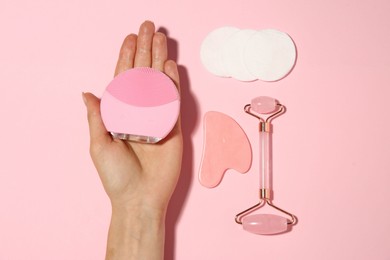 Woman holding facial cleansing brush near other skin care products on pink background, top view