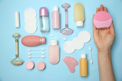 Woman holding facial cleansing brush near other body care products on light blue background, top view