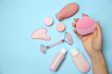 Woman holding facial cleansing brush near other skin care products on light blue background, top view. Space for text