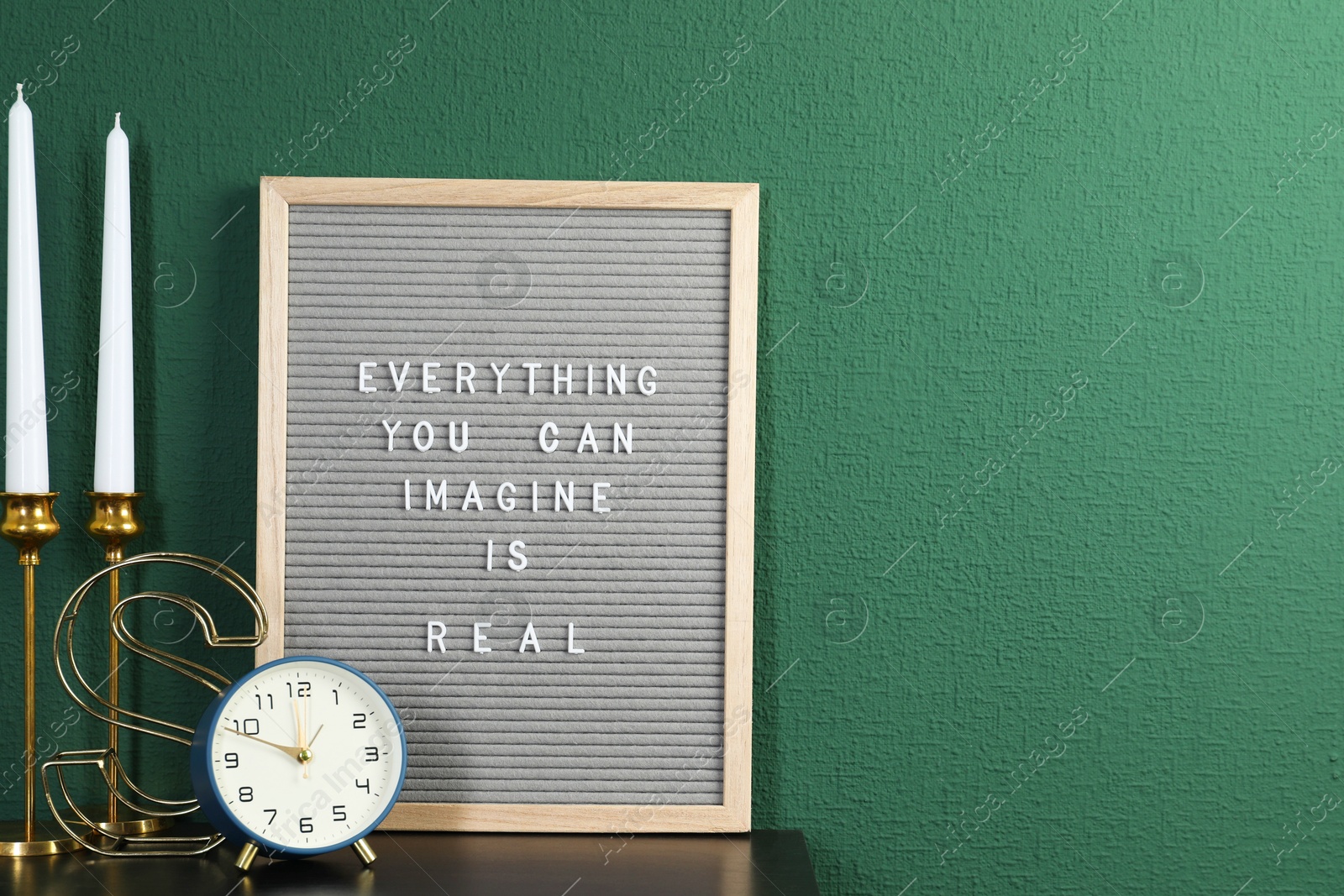 Photo of Letter board with phrase Everything You Can Imagine Is Real, alarm clock, candles and decor element on table near green wall. Space for text