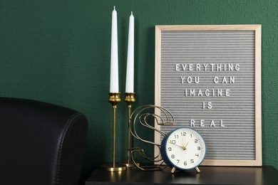 Letter board with phrase Everything You Can Imagine Is Real, alarm clock, candles and decor element on table near green wall