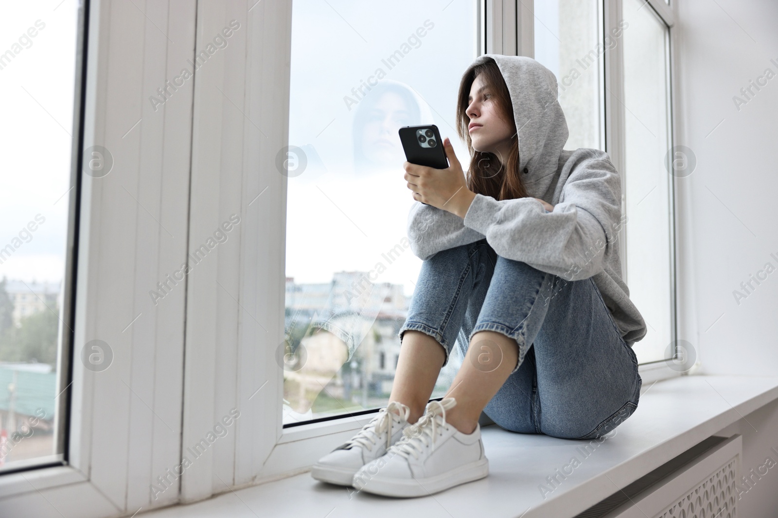 Photo of Loneliness concept. Sad teenage girl using smartphone on windowsill at home