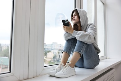 Loneliness concept. Sad teenage girl using smartphone on windowsill at home