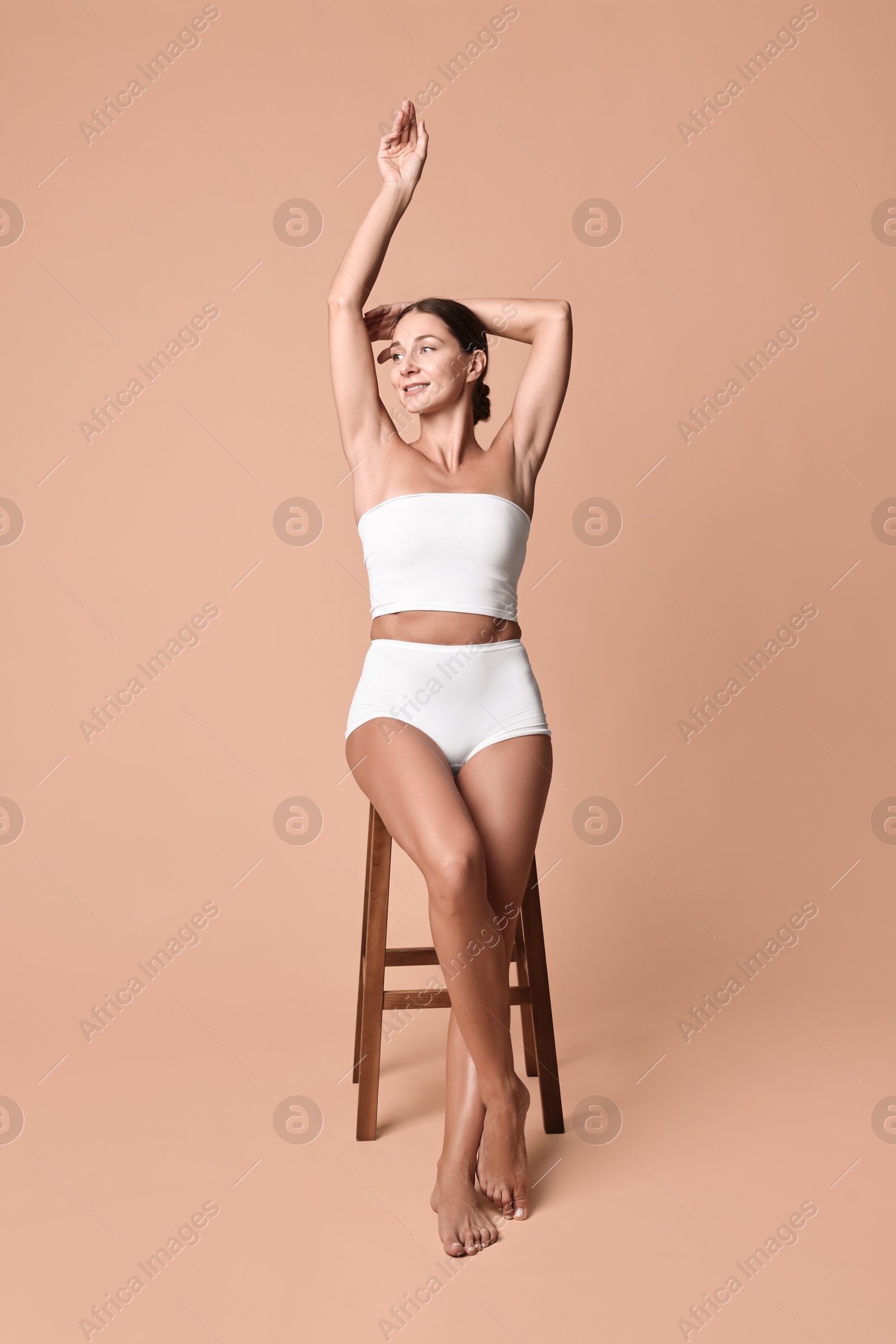 Photo of Smiling woman with perfect skin in underwear posing on stool against beige background. Body care