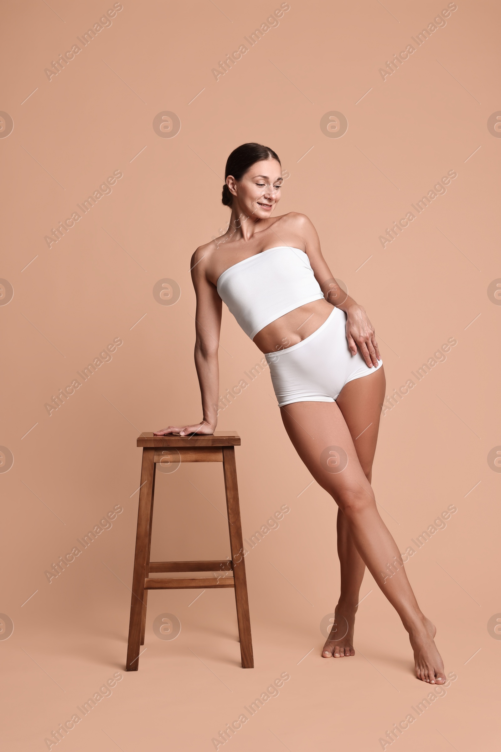 Photo of Smiling woman with perfect skin in underwear posing with stool against beige background. Body care