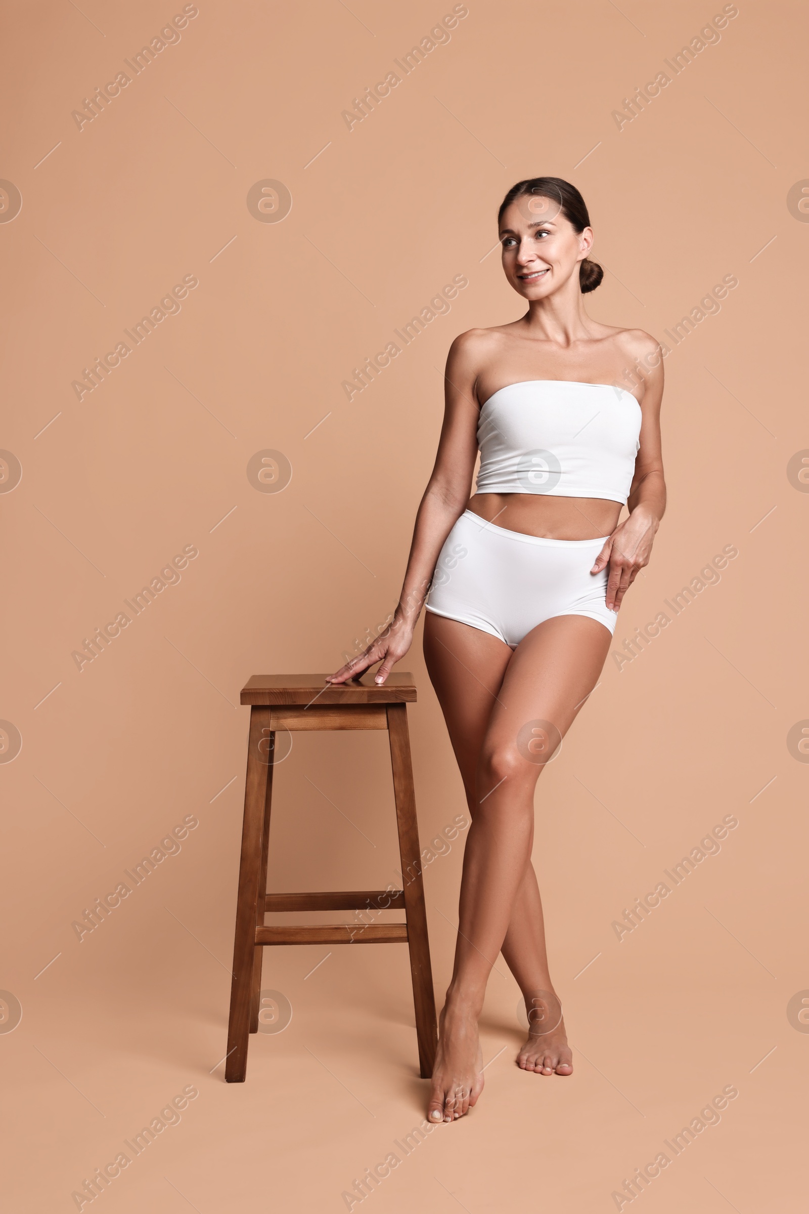 Photo of Smiling woman with perfect skin in underwear posing with stool against beige background. Body care