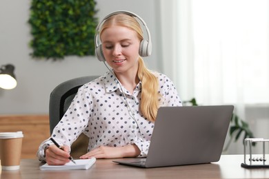 Interpreter in headphones taking notes while working with laptop at table indoors