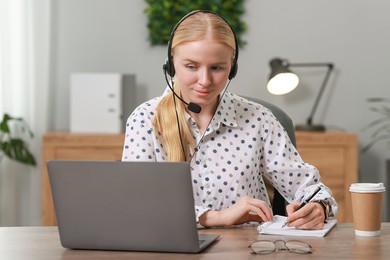 Interpreter in headset taking notes while working with laptop at table indoors