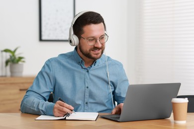 Interpreter in headphones taking notes while having video chat via laptop at wooden table indoors