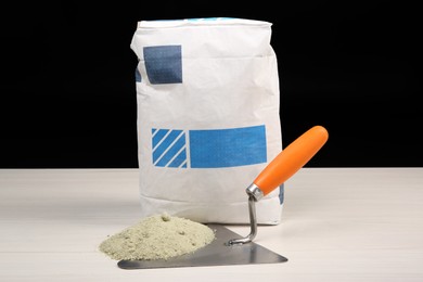 Photo of Bag of cement and putty knife on light wooden table against black background. Building material