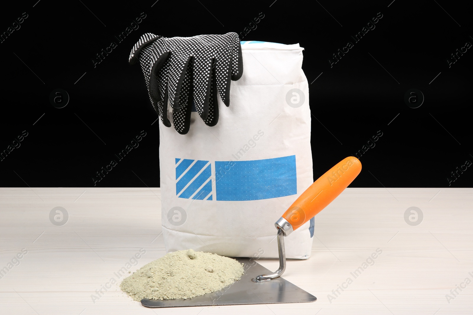 Photo of Bag of cement and putty knife on light wooden table against black background. Building material