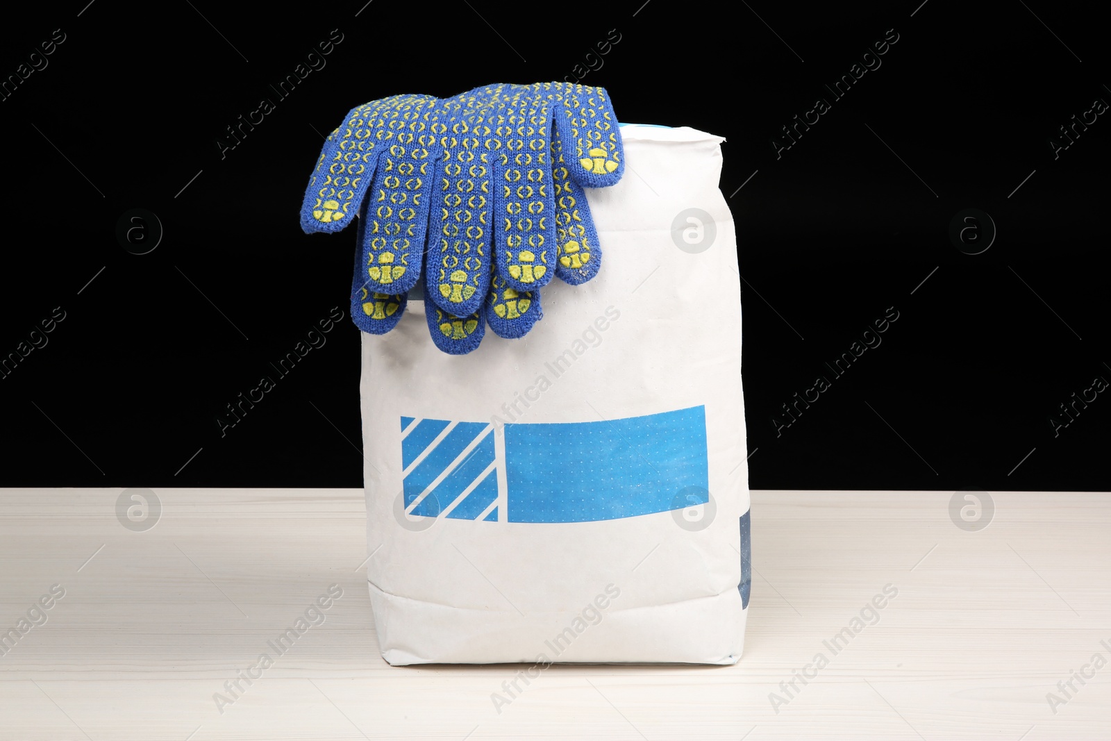 Photo of Bag of cement powder and rubber gloves on light wooden table against black background. Building material