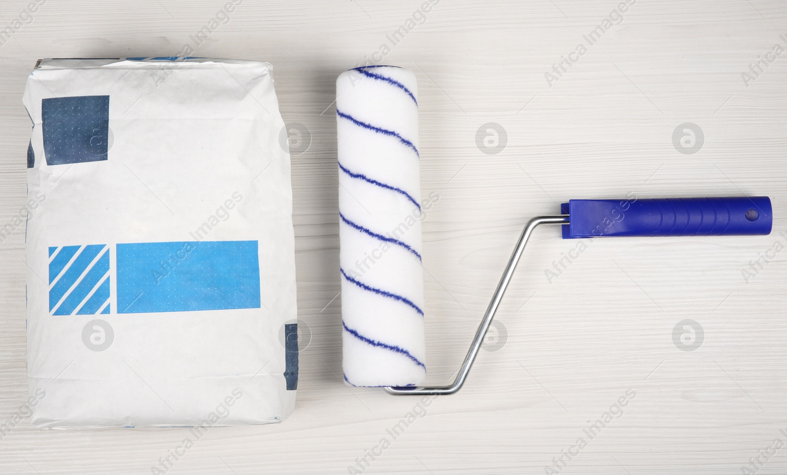 Photo of Bag of cement powder, and paint roller on light wooden table, flat lay. Building material