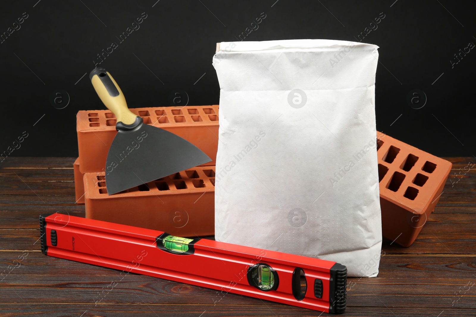 Photo of Red bricks, bag of cement powder, putty knife and spirit level on wooden table. Building materials and construction tools