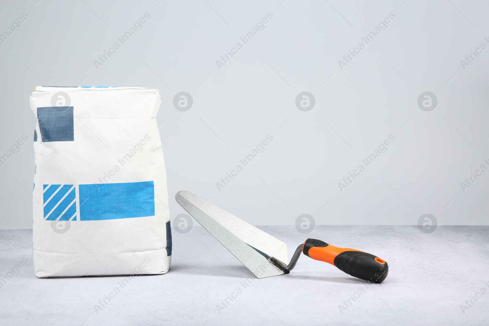 Photo of Bag of cement and trowel on textured table against light background, space for text. Building material