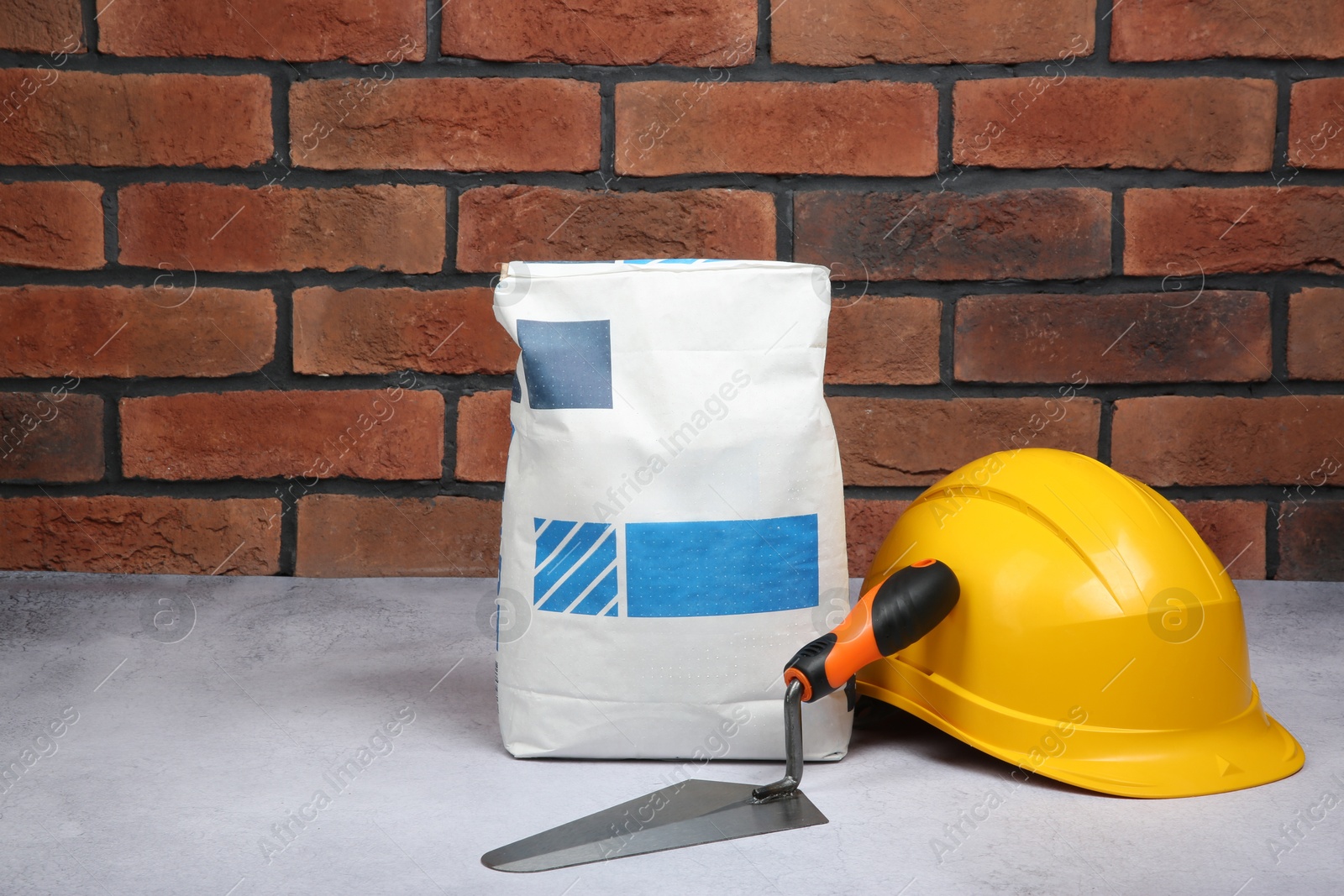 Photo of Bag of cement, hard hat and trowel on light textured table against red brick wall. Space for text