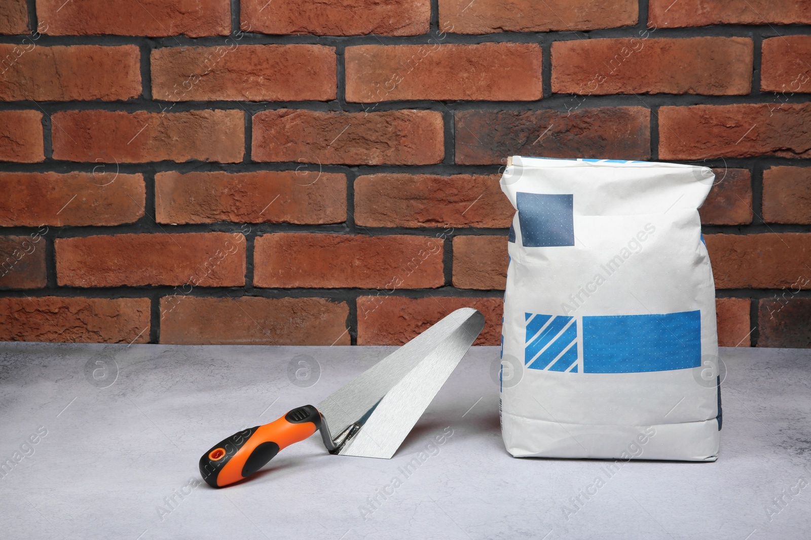 Photo of Bag of cement and trowel on light textured table against red brick wall. Space for text