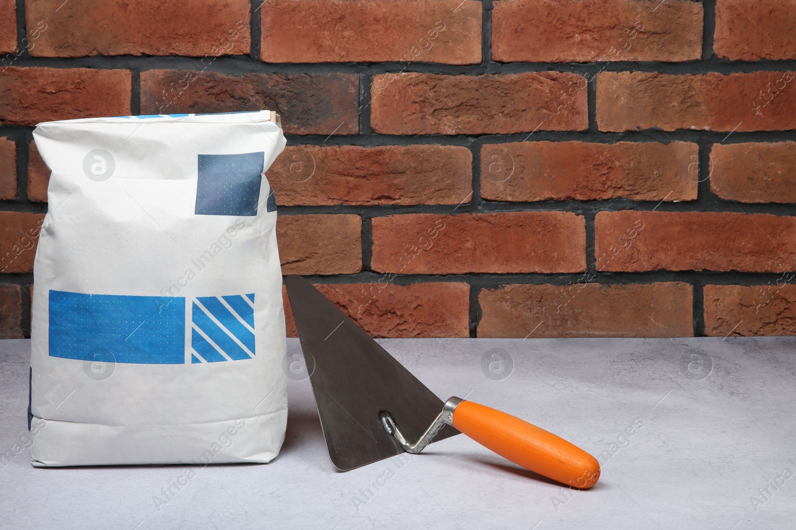 Photo of Bag of cement and trowel on light textured table against red brick wall. Space for text