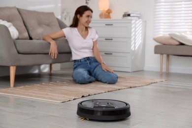 Photo of Young woman sitting in room, focus on robotic vacuum cleaner