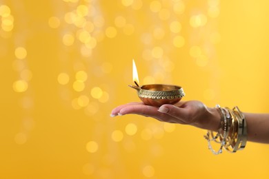 Photo of Diwali celebration. Woman holding lit diya lamp on yellow background with blurred lights, closeup. Space for text