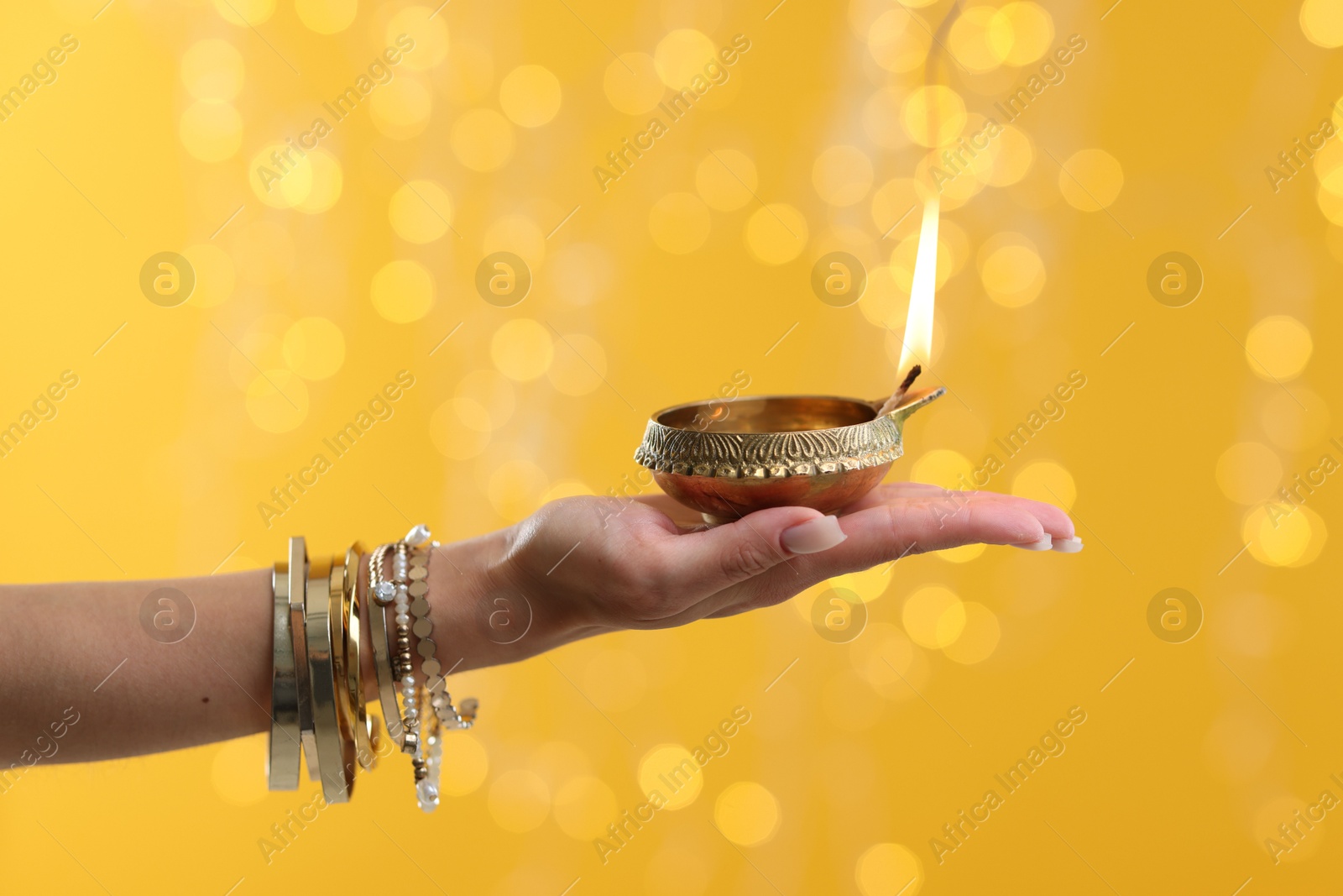 Photo of Diwali celebration. Woman holding lit diya lamp on yellow background with blurred lights, closeup