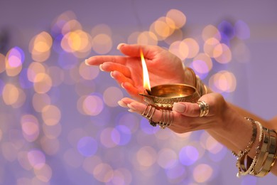 Photo of Diwali celebration. Woman holding lit diya lamp on color background with blurred lights, closeup