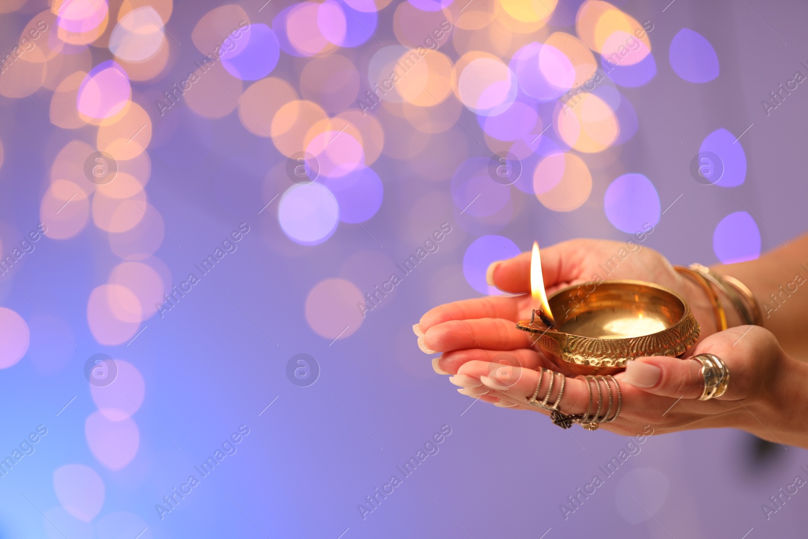 Photo of Diwali celebration. Woman holding lit diya lamp on color background with blurred lights, closeup. Space for text