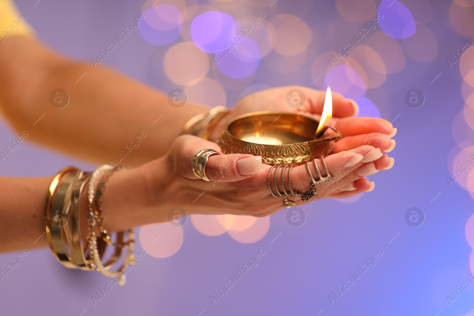 Photo of Diwali celebration. Woman holding lit diya lamp on color background with blurred lights, closeup