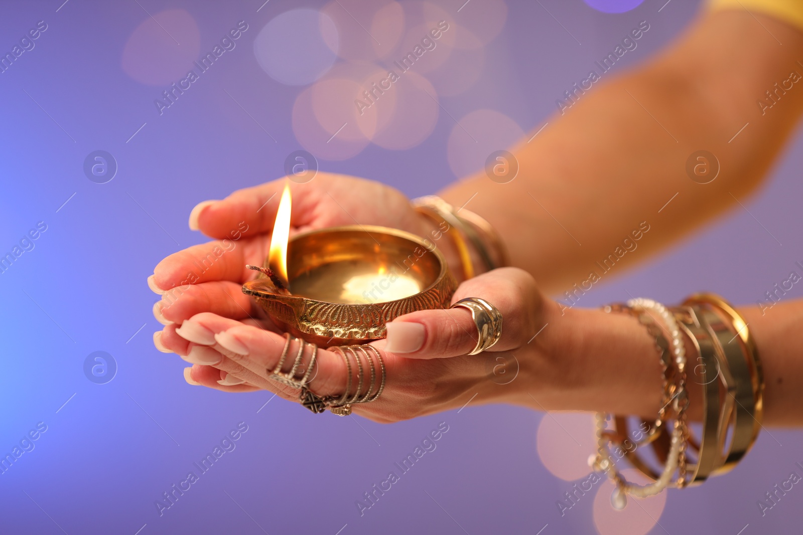 Photo of Diwali celebration. Woman holding lit diya lamp on color background with blurred lights, closeup