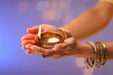 Photo of Diwali celebration. Woman holding lit diya lamp on color background with blurred lights, closeup