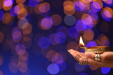 Diwali celebration. Woman holding lit diya lamp on color background with blurred lights, closeup. Space for text