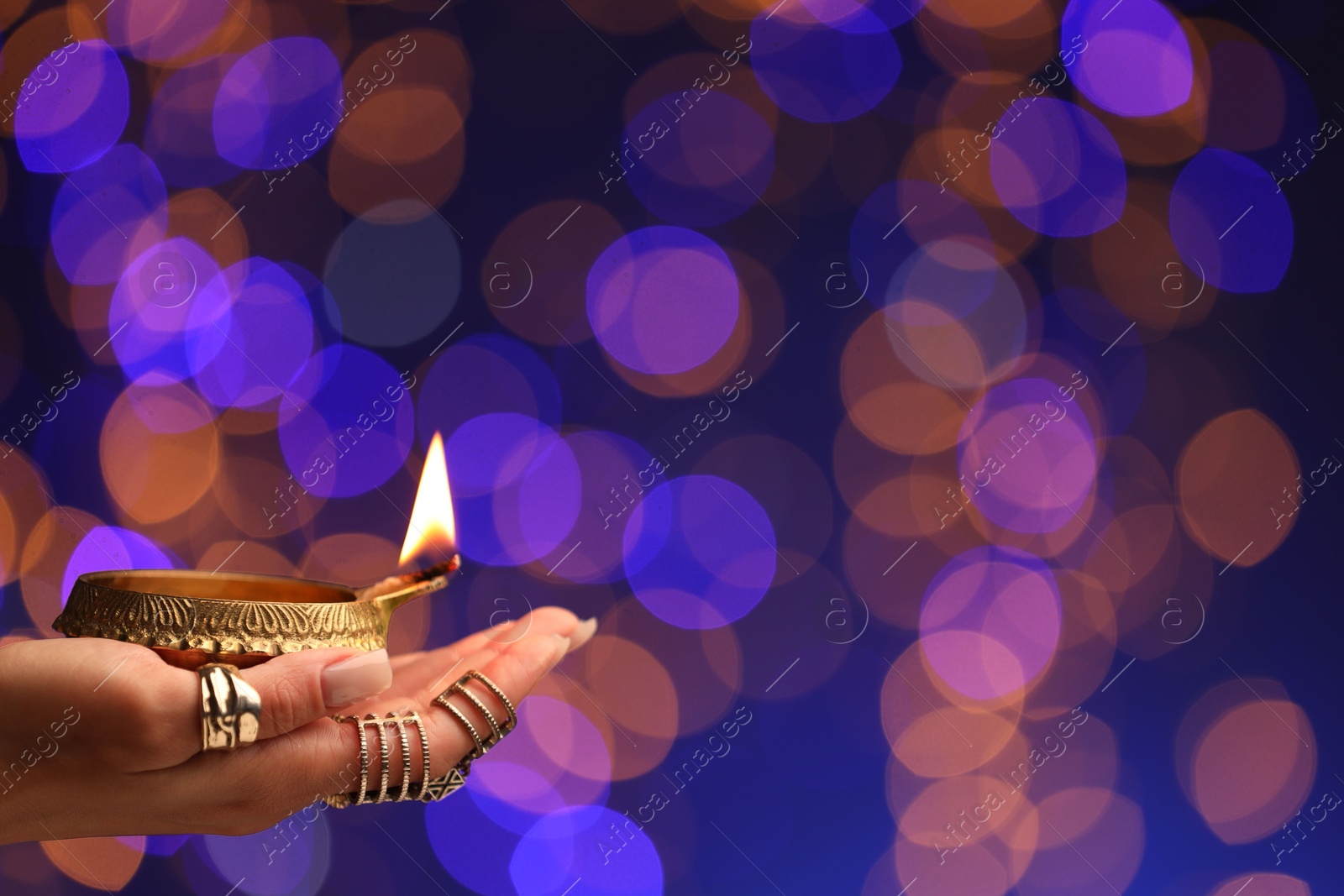 Photo of Diwali celebration. Woman holding lit diya lamp on color background with blurred lights, closeup. Space for text